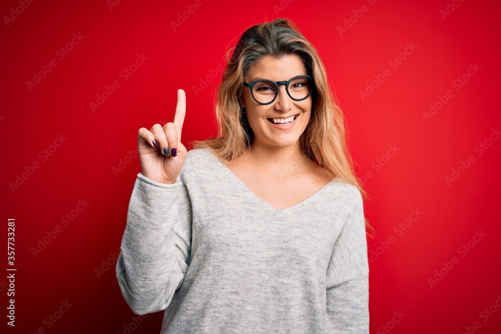 Young beautiful blonde woman wearing sweater and glasses over isolated red background showing and pointing up with finger number one while smiling confident and happy. Young beautiful blonde woman wearing sweater and glasses over isolated red background showing and pointing up with finger number one while smiling confident and happy.