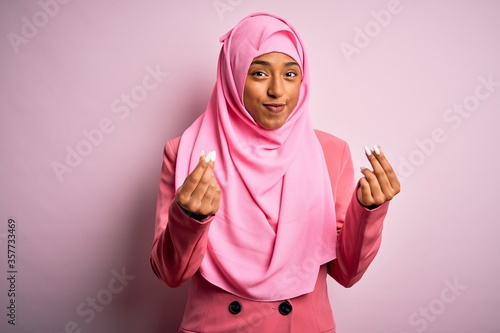 Young African American afro woman wearing muslim hijab over isolated pink background doing money gesture with hands, asking for salary payment, millionaire business © Krakenimages.com