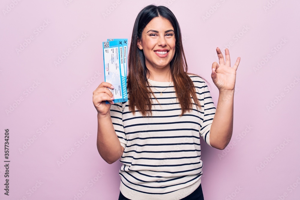 Young beautiful tourist woman holding airline boarding pass over isolated pink background doing ok sign with fingers, smiling friendly gesturing excellent symbol Young beautiful tourist woman holding airline boarding pass over isolated pink background doing ok sign with fingers, smiling friendly gesturing excellent symbol