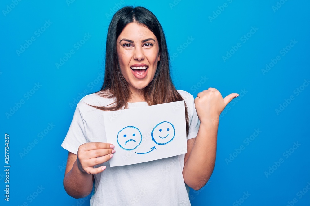 Woman asking for psychology change holding paper with unhappy to happy emotion message pointing thumb up to the side smiling happy with open mouth Woman asking for psychology change holding paper with unhappy to happy emotion message pointing thumb up to the side smiling happy with open mouth