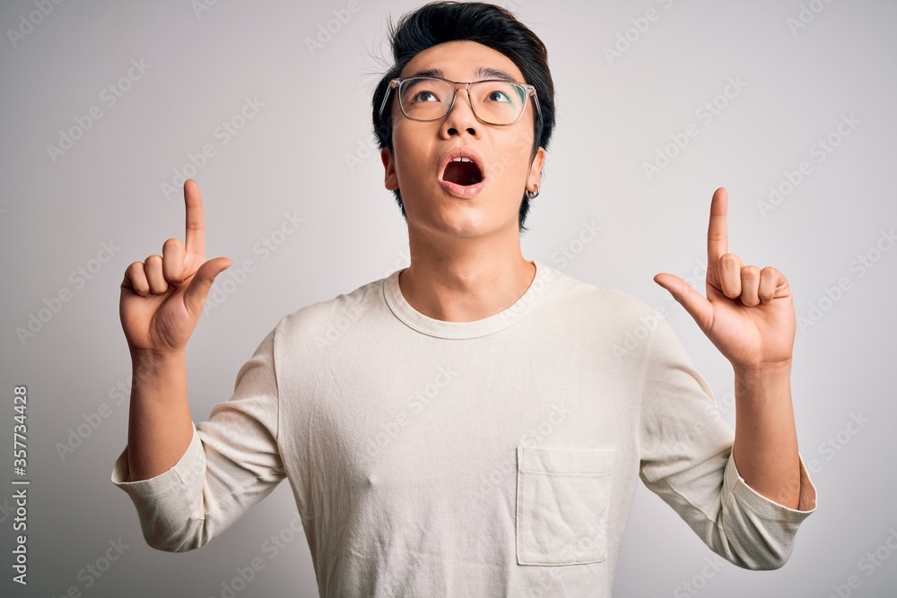 Young handsome chinese man wearing casual t-shirt and glasses over white background amazed and surprised looking up and pointing with fingers and raised arms. Young handsome chinese man wearing casual t-shirt and glasses over white background amazed and surprised looking up and pointing with fingers and raised arms.