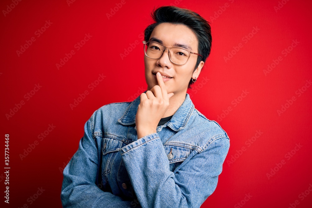 Young handsome chinese man wearing denim jacket and glasses over red background looking confident at the camera with smile with crossed arms and hand raised on chin. Thinking positive. Young handsome chinese man wearing denim jacket and glasses over red background looking confident at the camera with smile with crossed arms and hand raised on chin. Thinking positive.