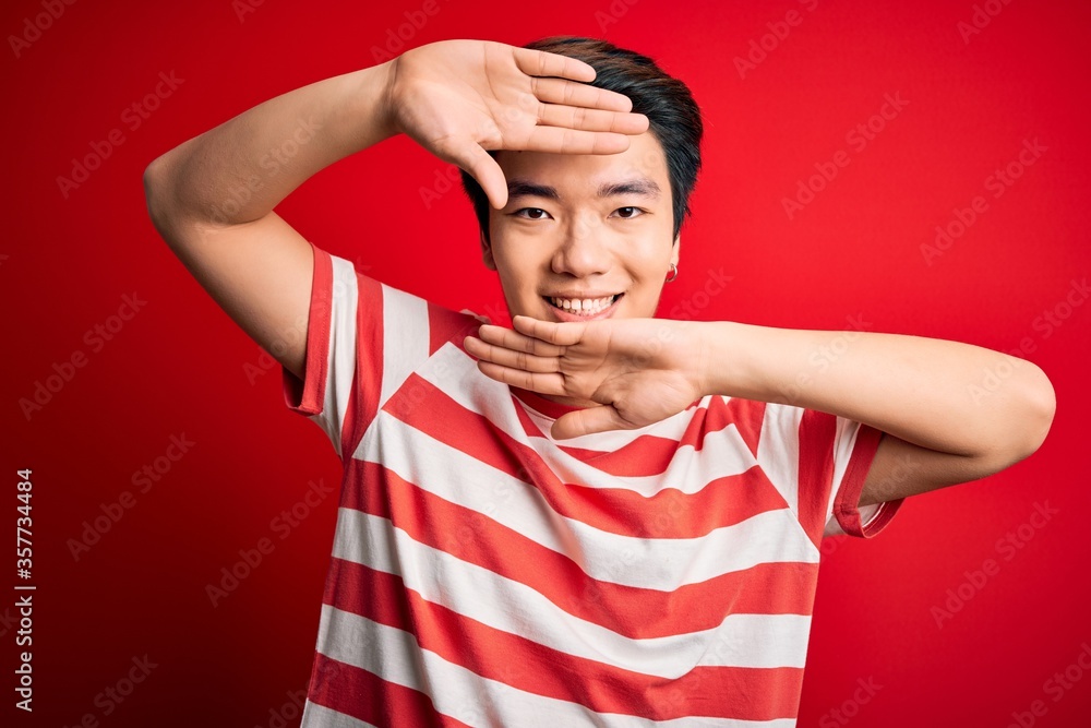 Fototapeta premium Young handsome chinese man wearing casual striped t-shirt standing over red background Smiling cheerful playing peek a boo with hands showing face. Surprised and exited