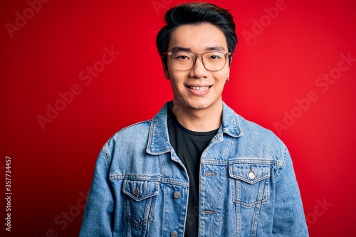 Young handsome chinese man wearing denim jacket and glasses over red background with a happy and cool smile on face. Lucky person.