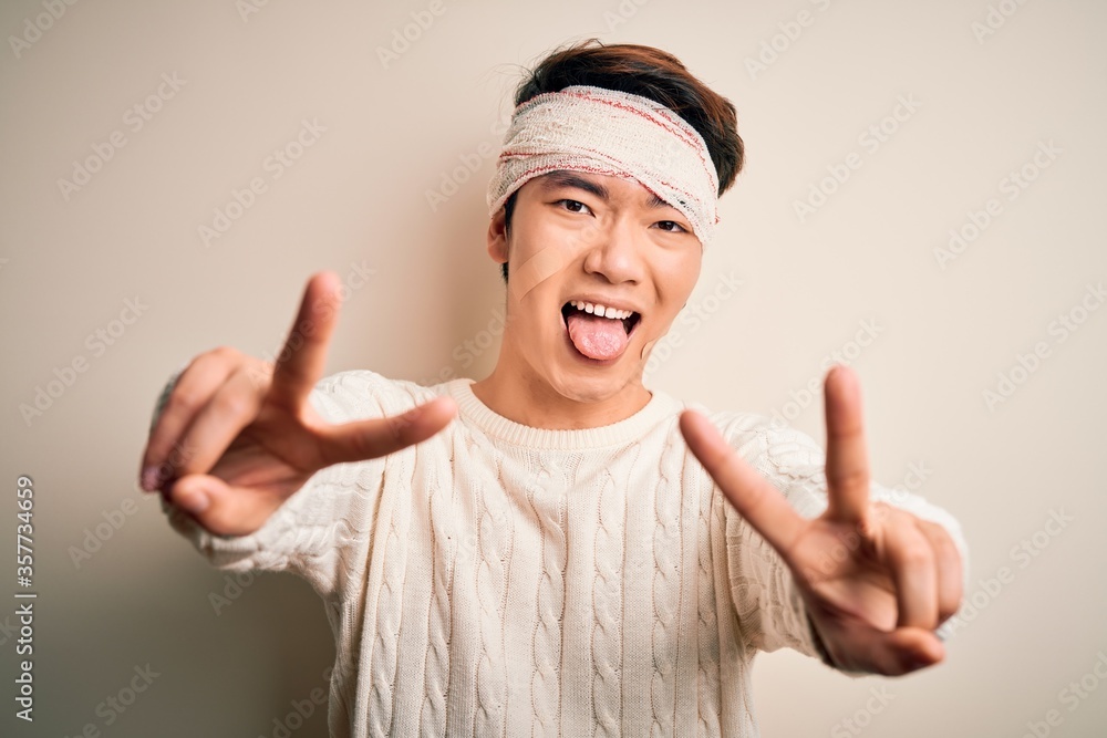 Young handsome chinese man injured for accident wearing bandage and strips on head smiling with tongue out showing fingers of both hands doing victory sign. Number two. Young handsome chinese man injured for accident wearing bandage and strips on head smiling with tongue out showing fingers of both hands doing victory sign. Number two.