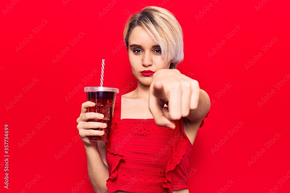 Young beautiful blonde woman drinking glass of cola beverage pointing with finger to the camera and to you, confident gesture looking serious Young beautiful blonde woman drinking glass of cola beverage pointing with finger to the camera and to you, confident gesture looking serious