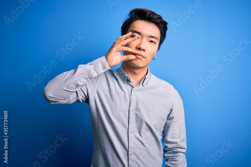 Young handsome chinese man wearing casual shirt standing over isolated blue background mouth and lips shut as zip with fingers. Secret and silent, taboo talking