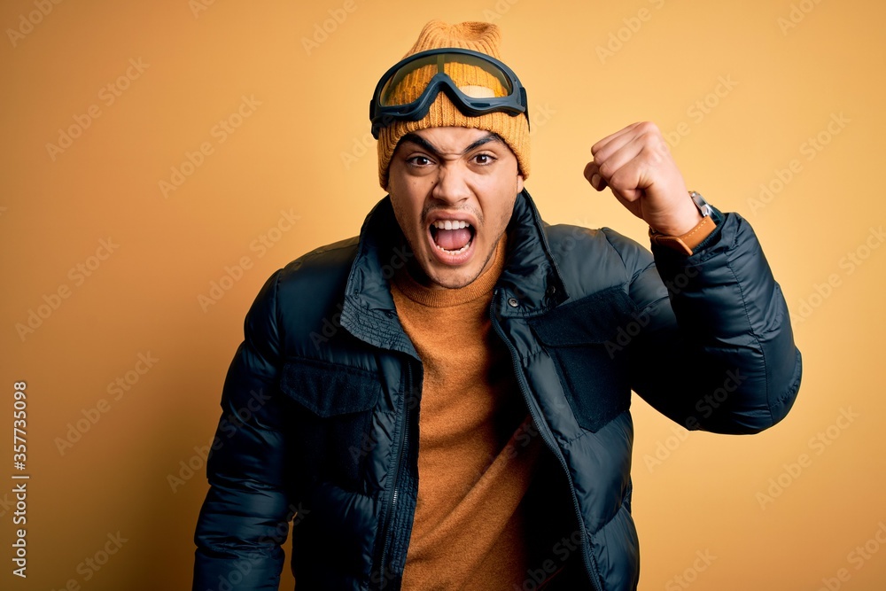 Young brazilian skier man wearing snow sportswear and ski goggles over yellow background angry and mad raising fist frustrated and furious while shouting with anger. Rage and aggressive concept. Young brazilian skier man wearing snow sportswear and ski goggles over yellow background angry and mad raising fist frustrated and furious while shouting with anger. Rage and aggressive concept.