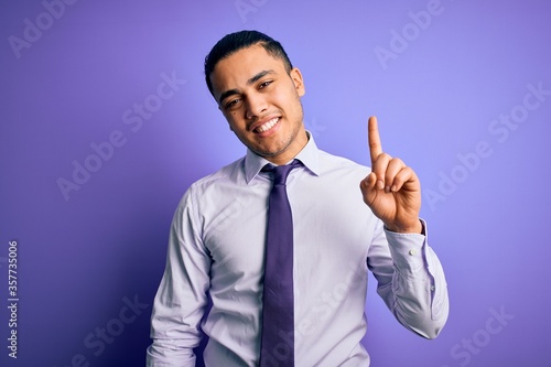 Young brazilian businessman wearing elegant tie standing over isolated purple background pointing finger up with successful idea. Exited and happy. Number one.