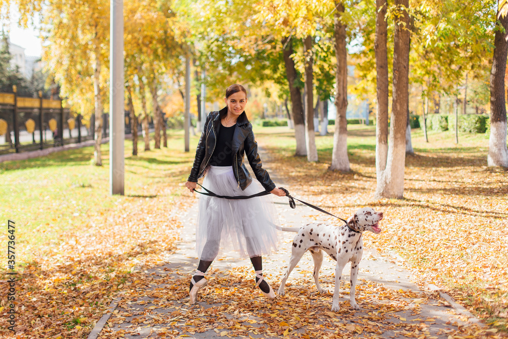 Fototapeta premium Ballerina with Dalmatian dog in the Park. Woman ballerina in a white ballet skirt and black leather jacket dancing in pointe shoes in autumn park with her spotty dalmatian dog.