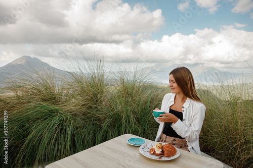 Young traveler woman sitting in outdoor cafe terrace with mountains view, drink coffee and eat croissant