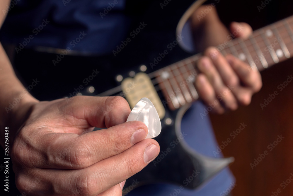 Closeup to a male hand holding a white guitar pick with a blurred black and yellow electric guitar at background. Rock & roll and music concept