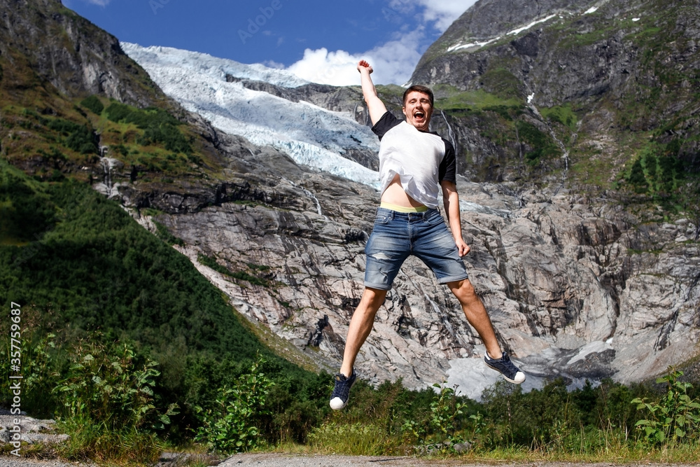 Trip to Norway. Young happy traveller is jumping on the background of melting glacier Boyabreen (Boya) from giant grey rocky mountain in the sunny summer day