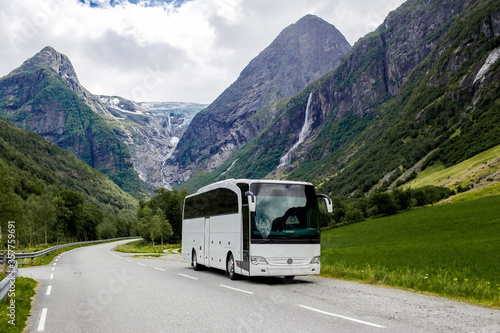 Blue ice tongue of Jostedal glacier melts from the giant rocky mountains into the green valley with waterfalls. Big white tourist bus rides on the road in Norway in summer cloudy day