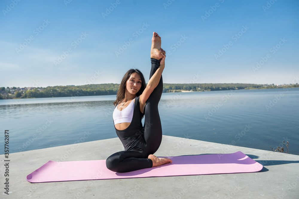 Fototapeta premium Sportive woman doing yoga exercise in the park, near the lake at daytime. Healthy lifestyle concept.