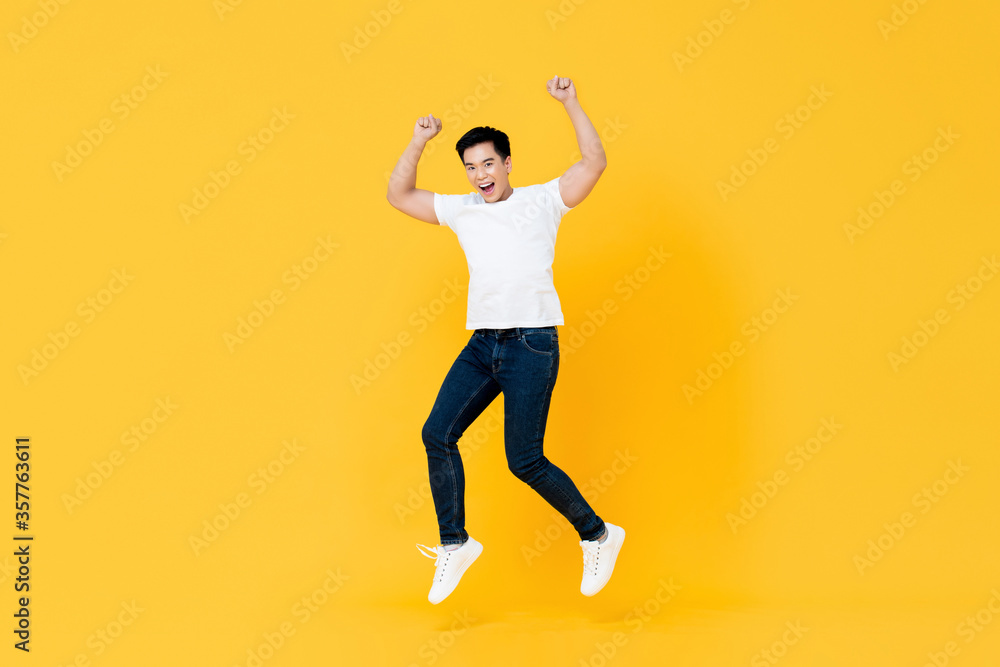 Happy young Asian man jumping with hands up isolated on yellow background