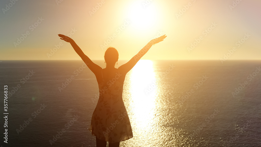 woman in summer dress silhouette raises hands standing on endless ocean beach edge against bright setting sun backside view