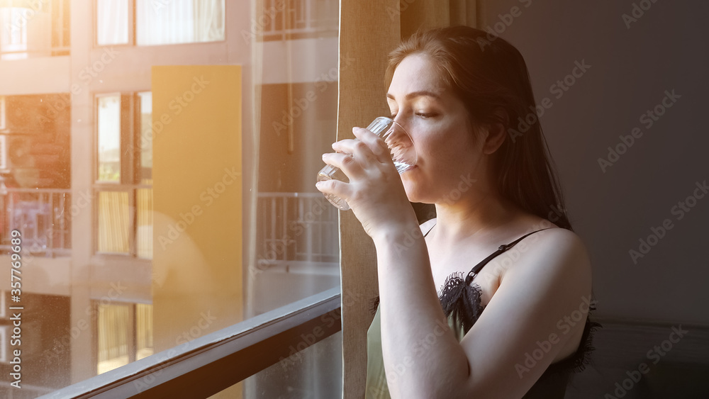 happy brunette lady in pajama drinks water from glass looking outside window at morning sunlight in hotel room closeup happy brunette lady in pajama drinks water from glass looking outside window at morning sunlight in hotel room closeup