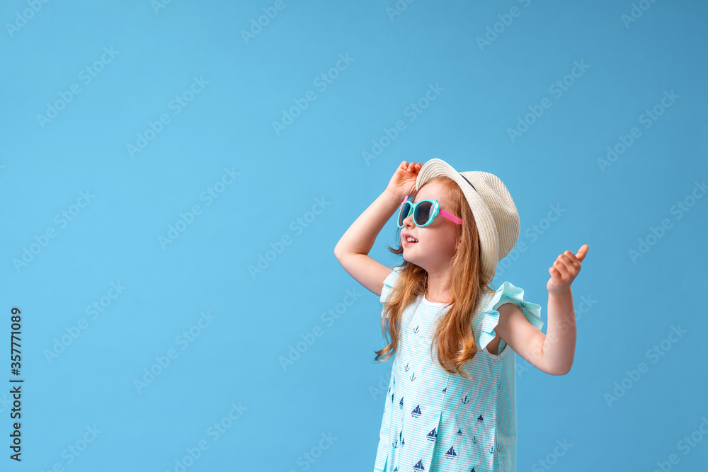 cute little girl in a dress, hat and sunglasses poses on a blue background