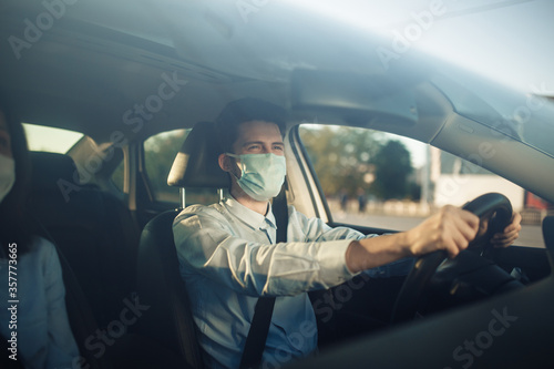 Young boy taxi driver holds his hands on the steering wheel and wears sterile medical mask. A man works during coronavirus pandemic and drives the passengers. Social distance and health care concept.