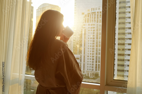 young pretty woman in bathrobe drinks coffee near window with city outside in apartment