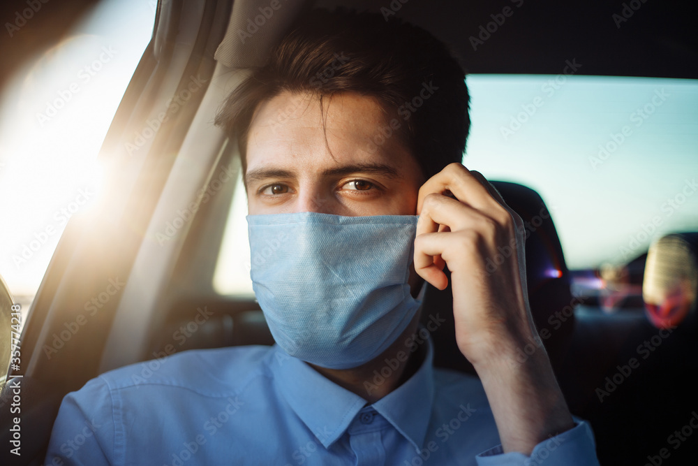 Young businessman takes a taxi and looks out of the car window adjusting sterile medical mask. Man sits on the back seat of taxi and takes a ride during coronavirus pandemic. Social distance concept. Young businessman takes a taxi and looks out of the car window adjusting sterile medical mask. Man sits on the back seat of taxi and takes a ride during coronavirus pandemic. Social distance concept.