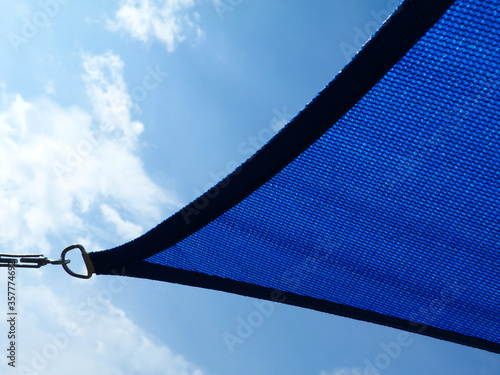 fabric sun shade and awning over patio or balcony. sun sail, spanning above city terrace. blue sky and white clouds. low angle view. metal connector and chain. summer and outdoors concept. photo