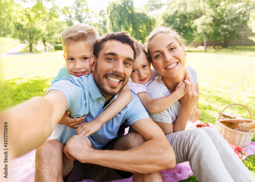 family, leisure and technology concept - happy mother, father and two little sons having picnic and taking selfie at summer park