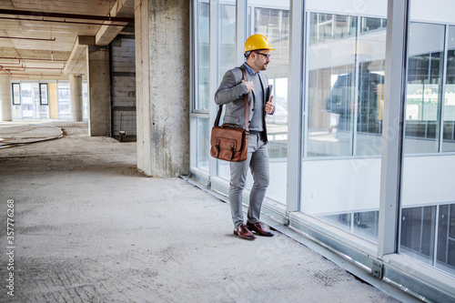 Young unshaven architect in suit with helmet on head standing in building in construction process and looking trough window. © dusanpetkovic1