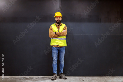 Young smiling attractive bearded worker in vest with helmet on head standing in front of black door with arms crossed and looking at camera.