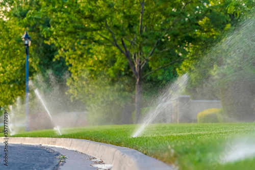 Paved road and curb against vibrant green grassy terrain with water sprinklers © Jason