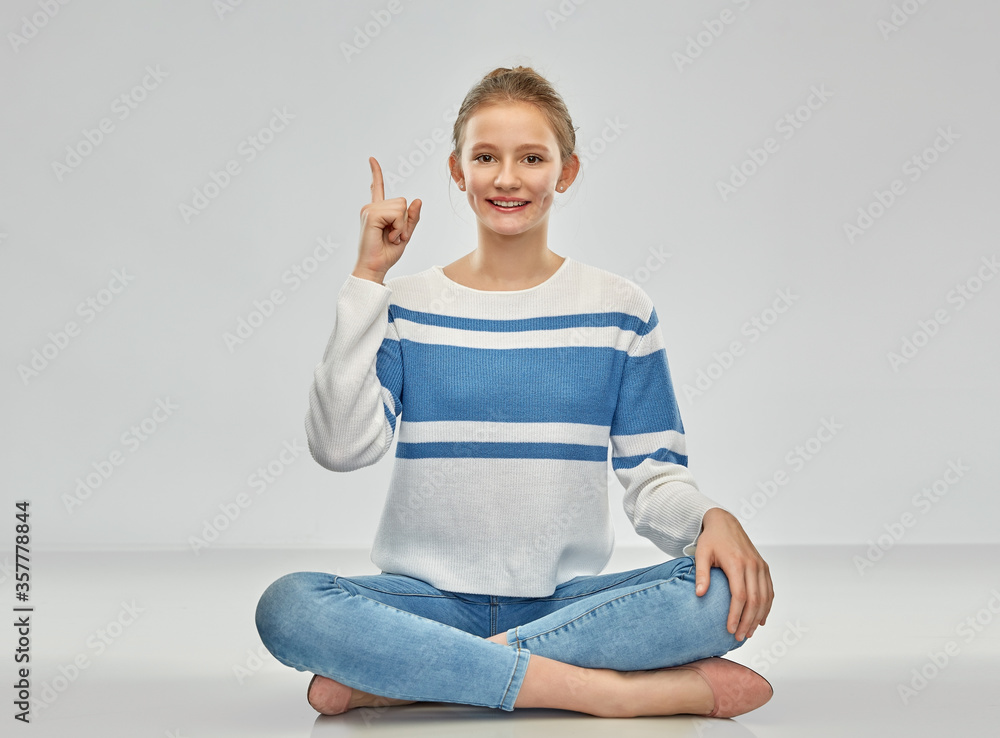 idea and people concept - smiling teenage girl in pullover pointing finger up sitting on floor over grey background idea and people concept - smiling teenage girl in pullover pointing finger up sitting on floor over grey background