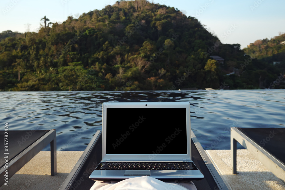 Fototapeta premium woman with laptop resting alone in lounge chair at hotel pool against forestry hill close back view