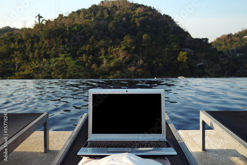 woman with laptop resting alone in lounge chair at hotel pool against forestry hill close back view