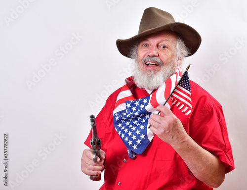 Smiling old cowboy with a pistol in one hand and the American flag in the other..Rancher is celebrating the 4th of July with a patriotic bandana, pistol and American flag.