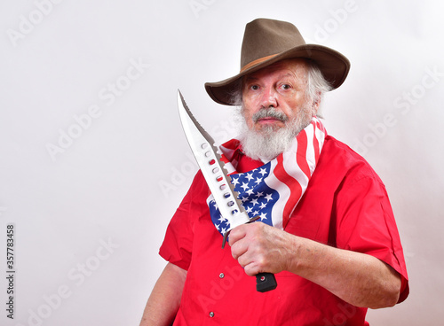 Bearded man wearing a patriotic bandana and a floppy western hat is showing off his giant hunting knfe..Cowvoy using his giant bowie knife to show off..