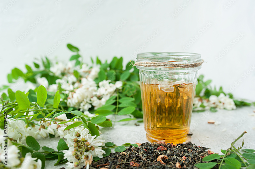 acacia flowers broth in glassware cup on a white cement background with lush flowering white acacia tree Branch , home self-treatment, useful properties of plants acacia flowers broth in glassware cup on a white cement background with lush flowering white acacia tree Branch , home self-treatment, useful properties of plants