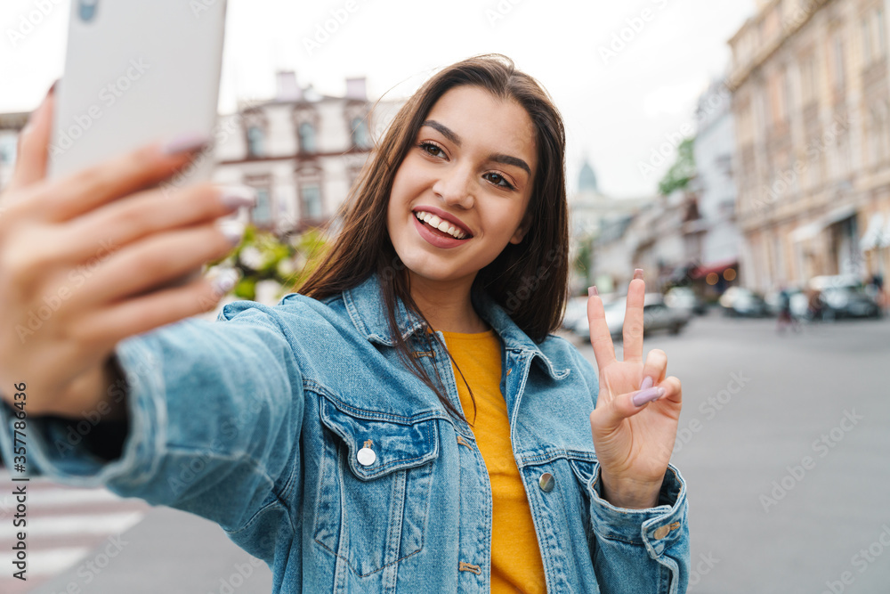 Image of woman taking selfie on cellphone and gesturing peace sign