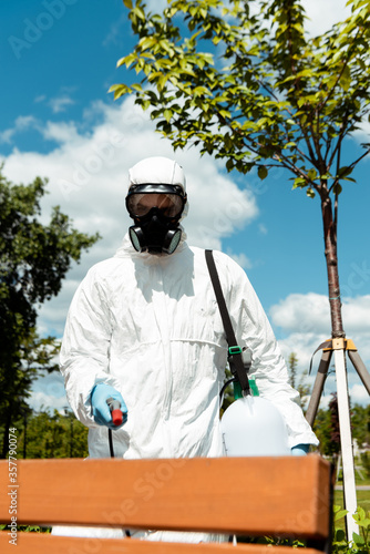 professional specialist in hazmat suit and respirator disinfecting bench in park during coronavirus pandemic