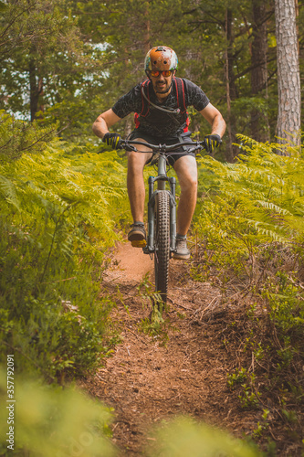 Man riding downhill with a mountain bike towards camera in a lush green environment on a dirt single track course. Tall man going downhill with a bike.