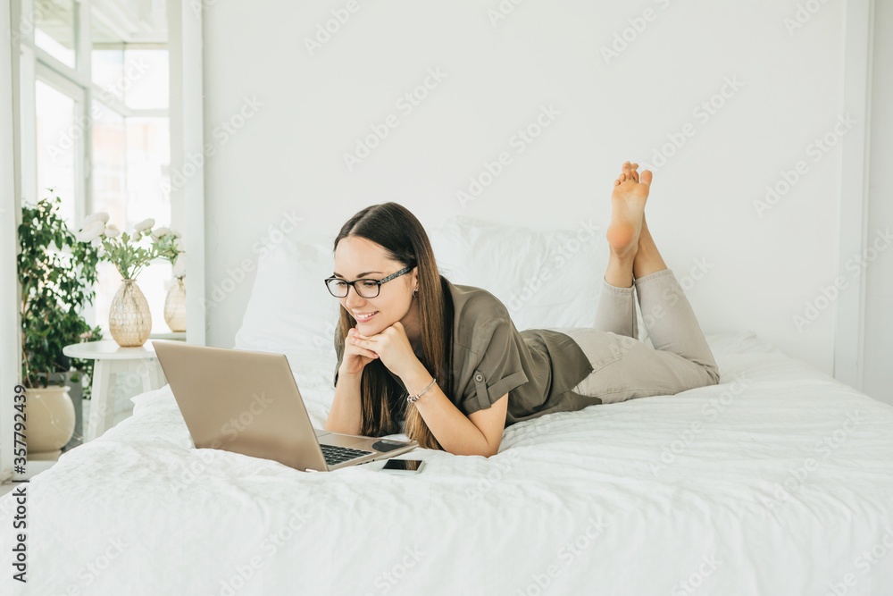 Positive girl student lies in bed and uses a laptop. She is reading from the screen or she is watching a video or training video course. Positive girl student lies in bed and uses a laptop. She is reading from the screen or she is watching a video or training video course.