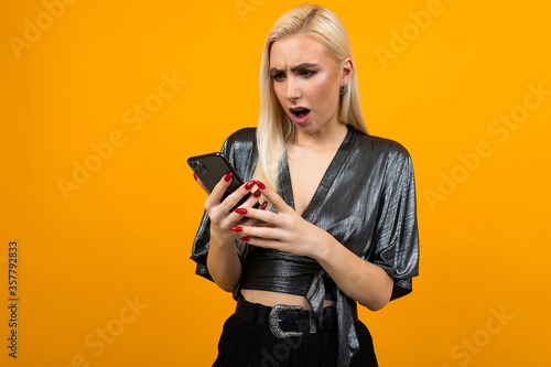 European stylish charming girl in surprise reads messages on her smartphone on a yellow studio background