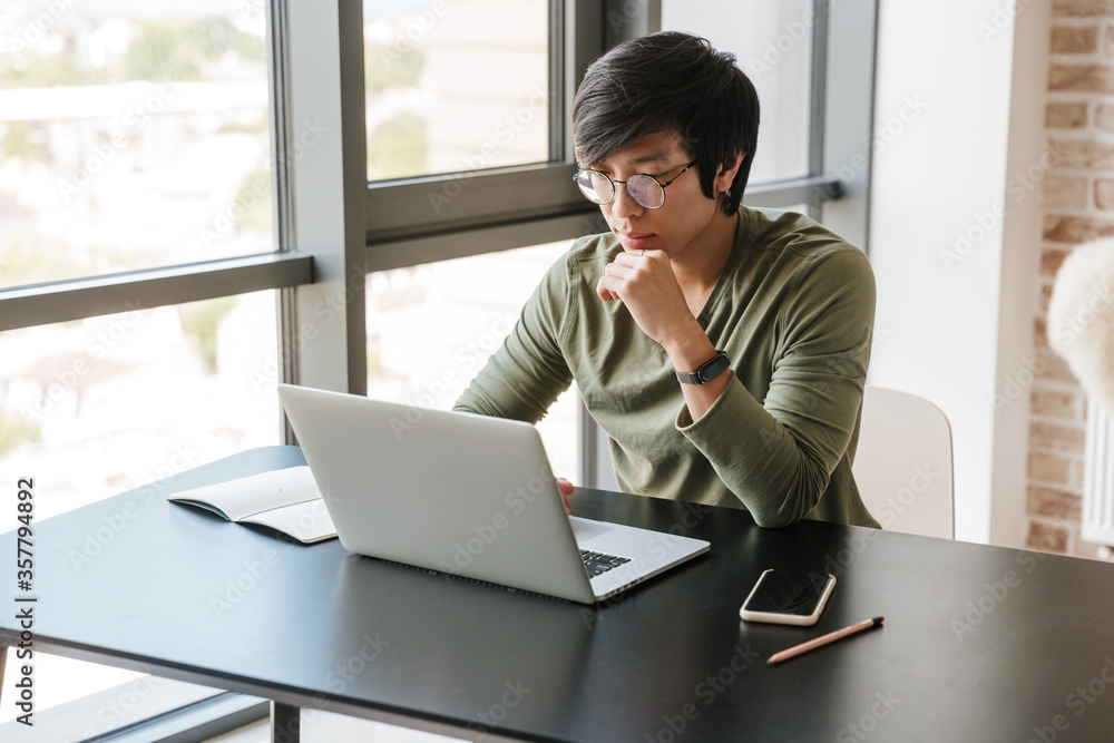 Image of serious asian man working with laptop while sitting at table Image of serious asian man working with laptop while sitting at table
