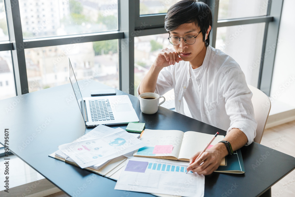 Image of focused asian man working with laptop and wireless earphones Image of focused asian man working with laptop and wireless earphones