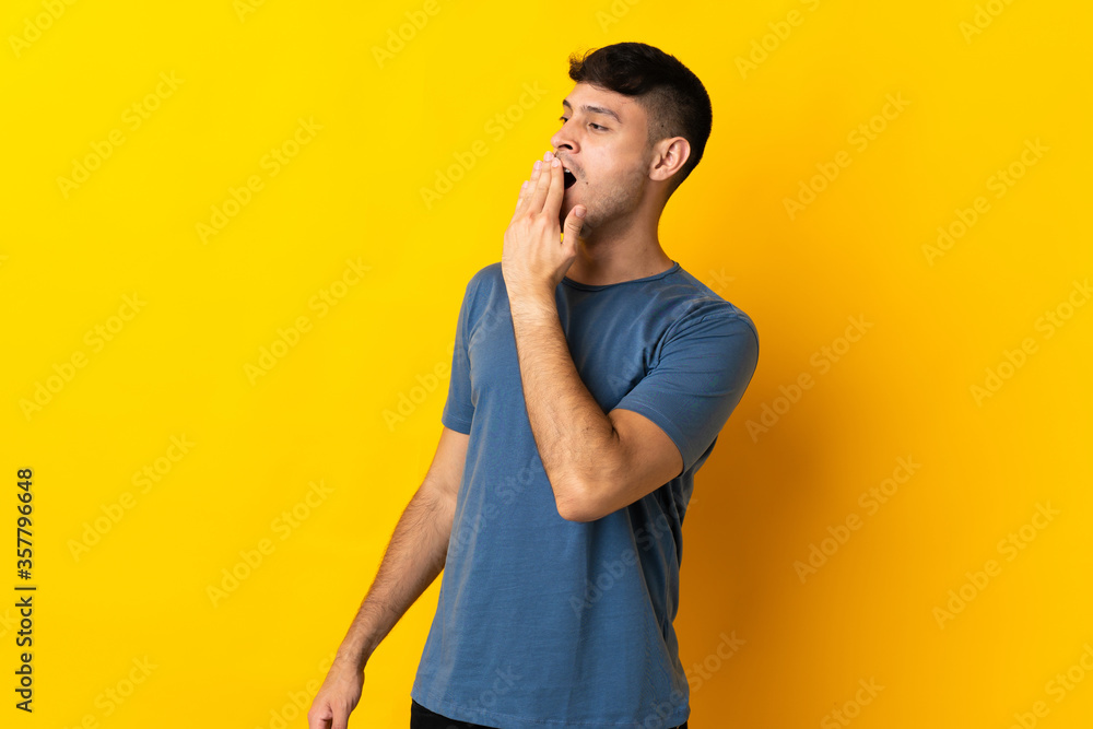 Young Colombian man isolated on yellow background yawning and covering wide open mouth with hand Young Colombian man isolated on yellow background yawning and covering wide open mouth with hand