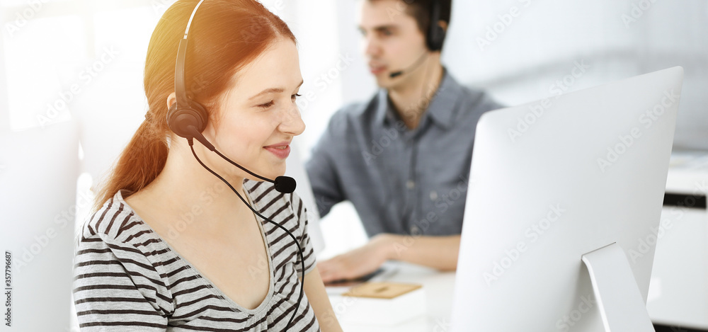 Casual dressed young woman using headset and computer while talking with customers online. Group of operators at work in sunny office. Call center Casual dressed young woman using headset and computer while talking with customers online. Group of operators at work in sunny office. Call center