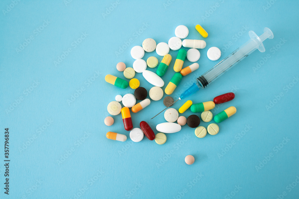 Multi-colored pills and capsules placed on a blue-green background with a syringe in the center of the conceptual image, isolated