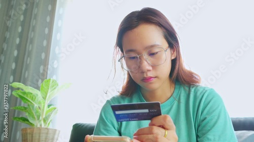 Young Asian woman sitting on a sofa holding a credit card while using a mobile device to type the credit card code Buy products online.
 photo