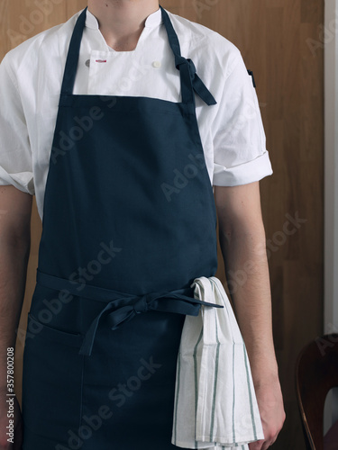 Slika na platnu A man cook in a blue apron and a white shirt on a wooden background, a towel in