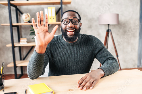 Excited African-American young guy in glasses looks into camera and greeting , waving hello. Video screen, video chat, online call, webcam shot photo